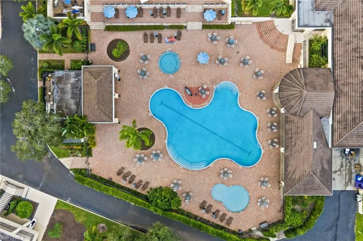 Aerial view of a sprawling pool complex featuring a large free-form swimming pool, a separate circular splash pool, a smaller secondary wading pool, extensive brick paver decking, and a covered architectural pavilion