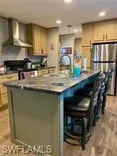Kitchen with wall chimney range hood, stainless steel refrigerator, a kitchen island with sink, and light hardwood / wood-style flooring