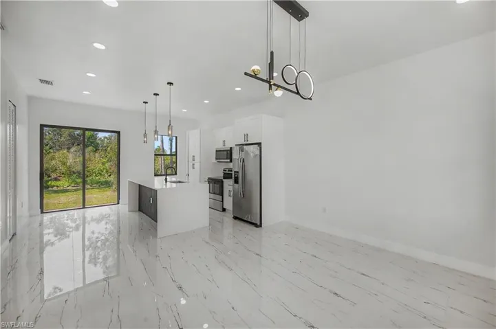 Modern kitchen featuring polished stone-finish flooring, stainless steel appliances, white cabinetry with waterfall-edge countertops, large sliding glass doors with natural light, and contemporary pendant lighting suspended from vaulted ceilings with recessed lighting throughout