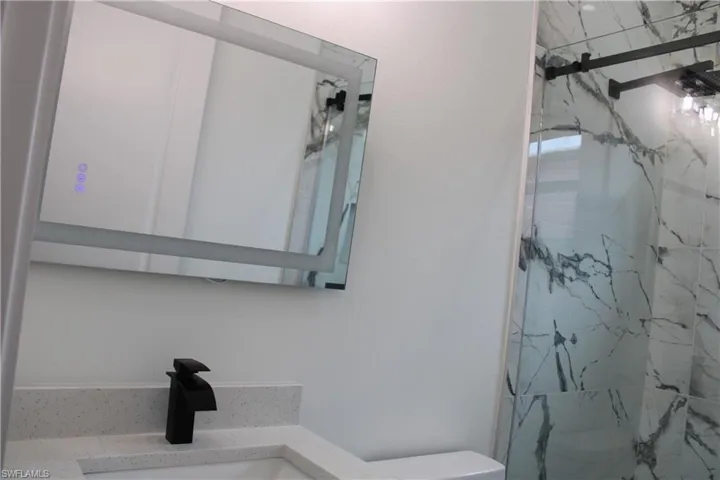 Bathroom featuring an illuminated mirror, black hardware, a light-toned countertop, and a shower with glass enclosure and veined tile surround