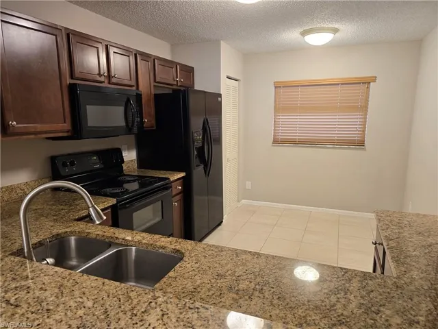 Kitchen with black appliances, a sink, a textured ceiling, light tile patterned floors, and light granite countertops