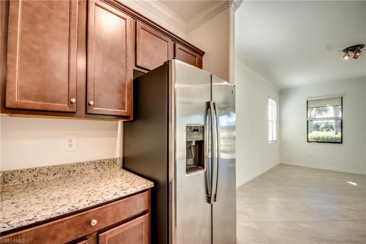 Kitchen featuring crown molding, stainless steel refrigerator with ice dispenser, light stone countertops, brown cabinetry, and light tile patterned floors