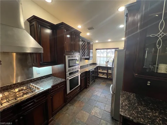 Kitchen featuring stone tile flooring, dark stone countertops, visible vents, wall chimney range hood, and appliances with stainless steel finishes