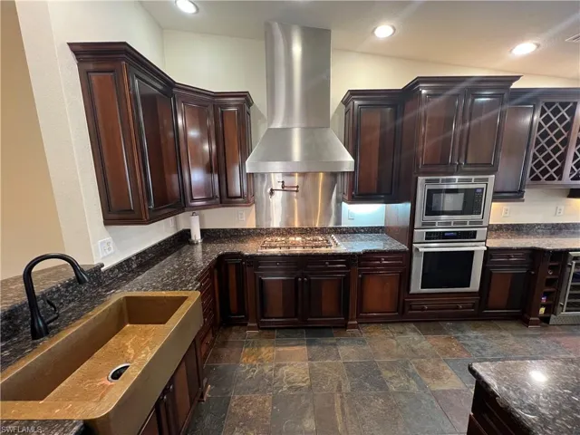 Kitchen with stainless steel appliances, dark stone countertops, wall chimney range hood, a sink, and stone finish flooring