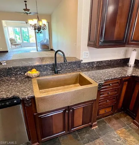 Kitchen with stainless steel dishwasher, a sink, stone finish flooring, dark stone counters, and a notable chandelier