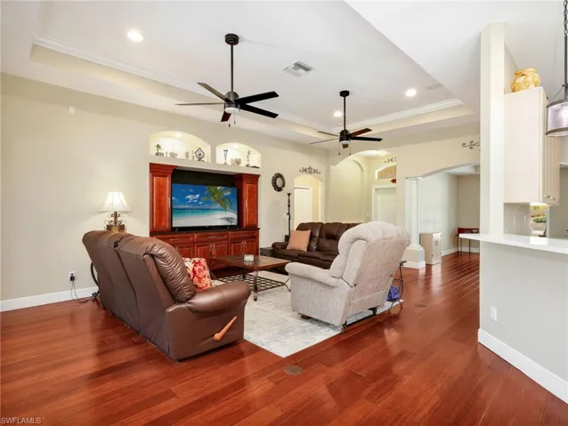 Living room with a tray ceiling, crown molding, arched walkways, dark wood-type flooring, and ornate columns