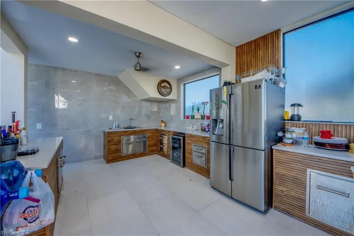 Kitchen with brown cabinets, stainless steel refrigerator with ice dispenser, tile walls, light stone countertops, and beverage cooler