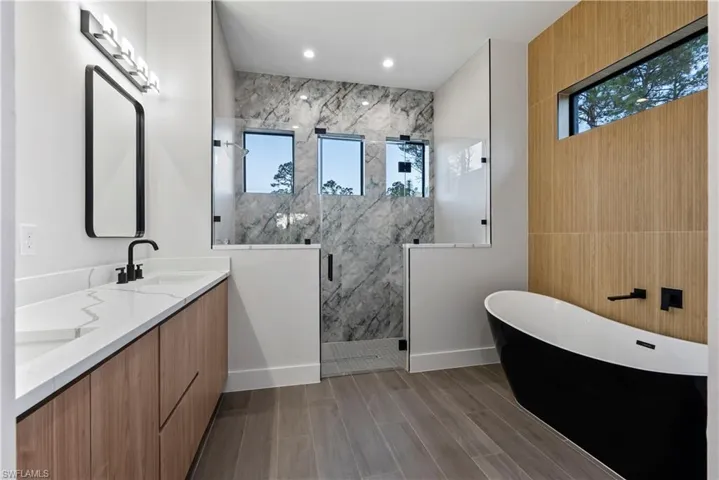 Modern bathroom featuring a freestanding black and white bathtub, a walk-in shower with clear glass enclosures and marble-look wall tiles, and a double vanity with wood-finish cabinetry and white countertops