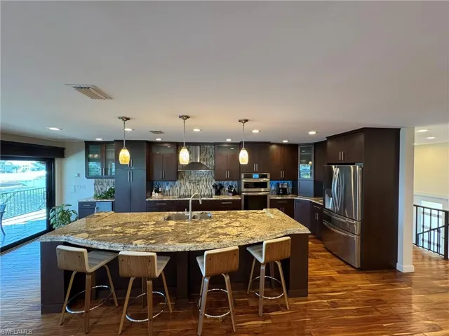 Kitchen featuring dark brown cabinets, pendant lighting, stainless steel appliances, a kitchen island with sink, and wall chimney range hood