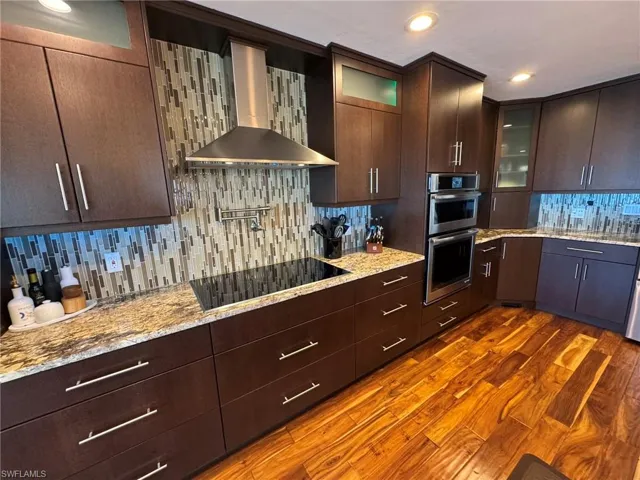 Kitchen featuring wall chimney range hood, double oven, black electric stovetop, light stone countertops, and dark hardwood / wood-style flooring