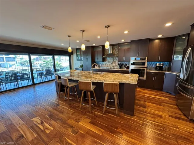 Kitchen featuring appliances with stainless steel finishes, decorative light fixtures, wall chimney range hood, dark brown cabinets, and a center island with sink