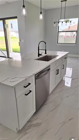 Kitchen featuring stainless steel dishwasher, a sink, light marble finish floors, decorative light fixtures, and white cabinets