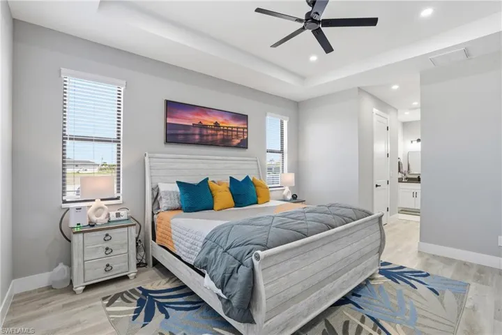 Bedroom with a raised ceiling, light wood-style floors, ensuite bath, ceiling fan, and recessed lighting