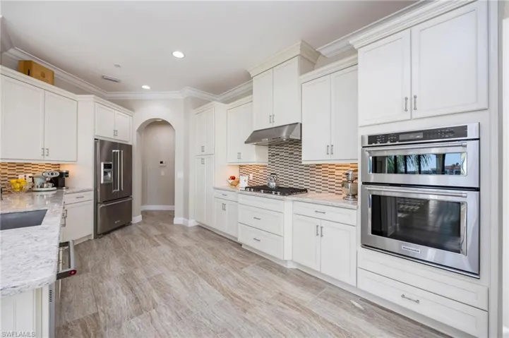 Kitchen featuring arched walkways, stainless steel appliances, white cabinets, light stone countertops, and ornamental molding