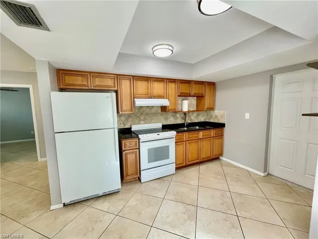 Kitchen featuring natural wood cabinetry, black countertops, tile backsplash, tile flooring, and white appliances including a refrigerator and range