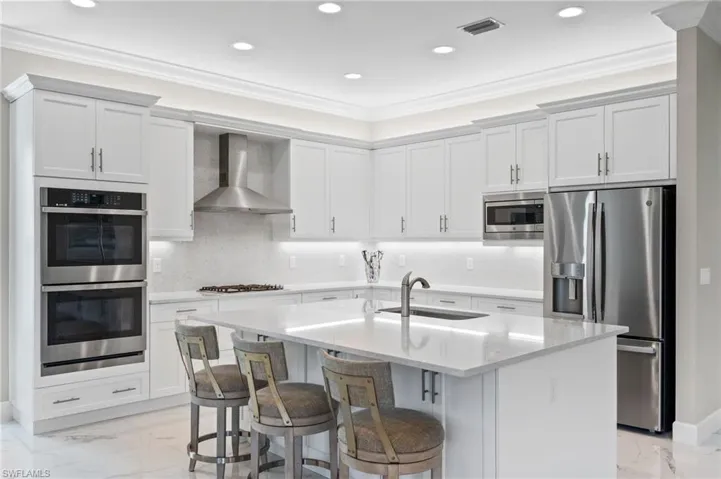 Kitchen with an island with sink, appliances with stainless steel finishes, ornamental molding, wall chimney exhaust hood, and sink