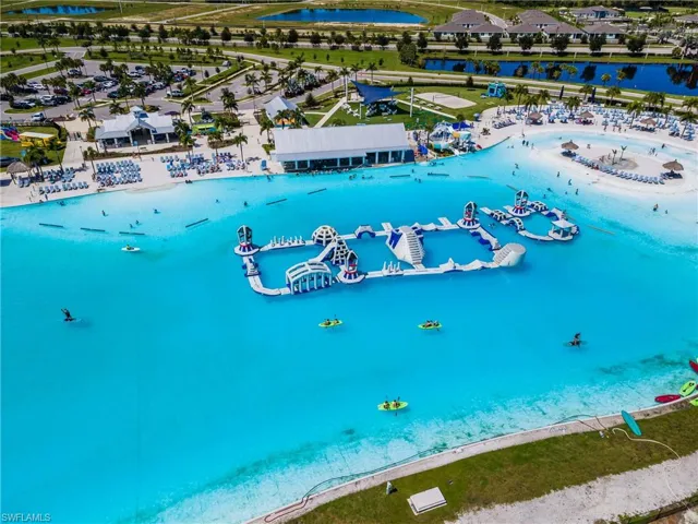 Expansive lagoon with clear blue water, large inflatable water obstacle course, sandy beach areas with lounge chairs, and surrounding recreational buildings