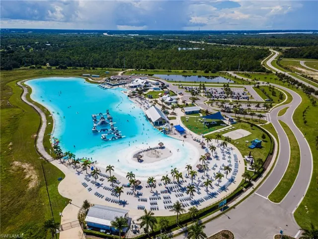 Expansive lagoon with a sandy beach and water features, surrounded by multiple recreation buildings, paved pathways, and mature palm trees