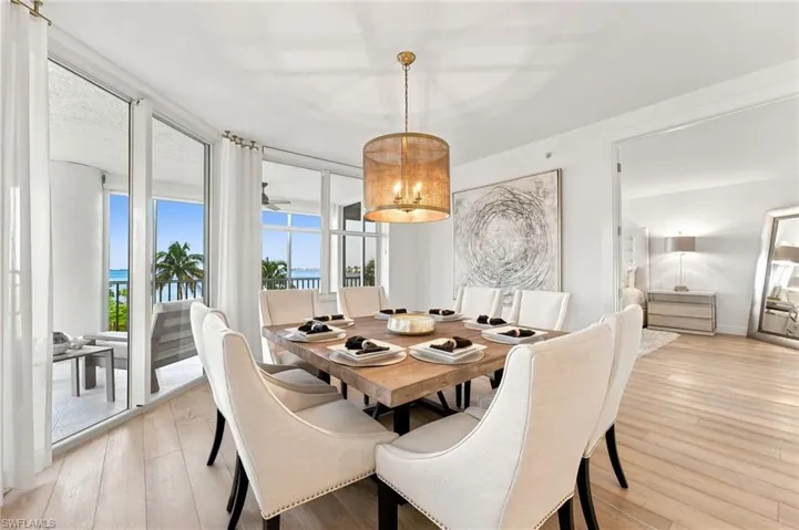 Open-concept dining area with wood-finish flooring, a contemporary chandelier, and a wide balcony with water and palm tree scenery