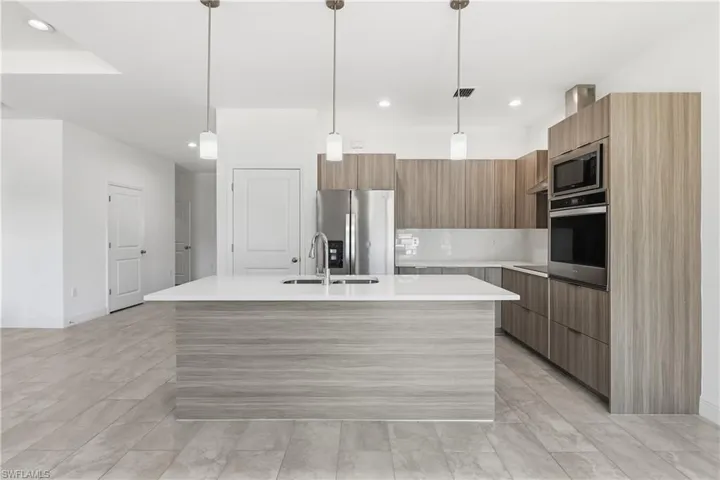 Kitchen with modern cabinets, an island with sink, hanging light fixtures, stainless steel appliances, and light stone counters