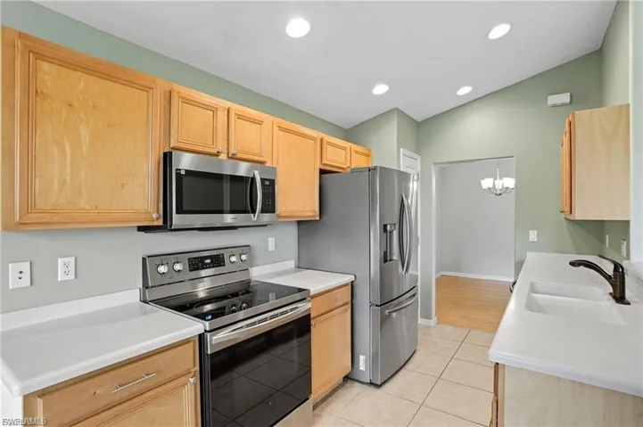 Kitchen with stainless steel appliances, light wood finish cabinets, a chandelier, light tile patterned flooring, and vaulted ceiling