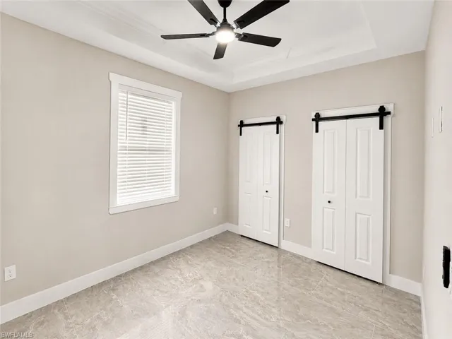 Room featuring modern tile flooring, two barn-door style closets, a ceiling fan with light fixture, a tray ceiling, and a window with blinds
