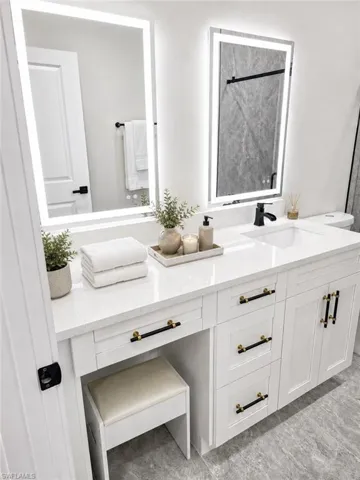 Spacious vanity featuring double illuminated mirrors, a white countertop, recessed sink, and black and gold hardware