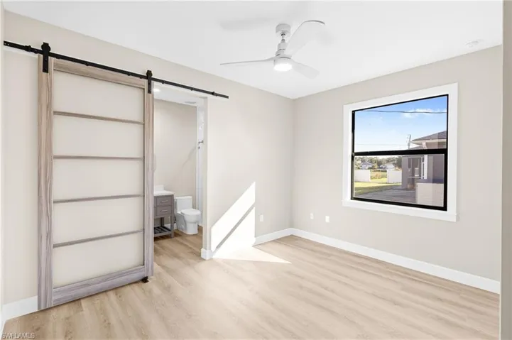 Unfurnished bedroom with a barn door, ensuite bathroom, light wood-type flooring, and ceiling fan
