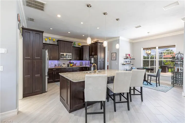 Kitchen featuring stainless steel appliances, dark brown cabinets, ornamental molding, light stone counters, and an island with sink