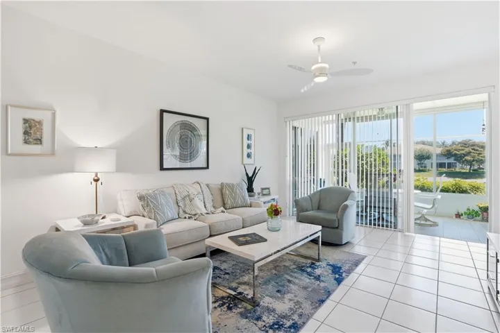 Bright living space with white tile flooring and a patio featuring a white railing, accessible via a large glass sliding door with vertical blinds
