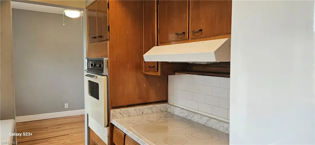 Kitchen with brown cabinets, decorative backsplash, light countertops, under cabinet range hood, and oven