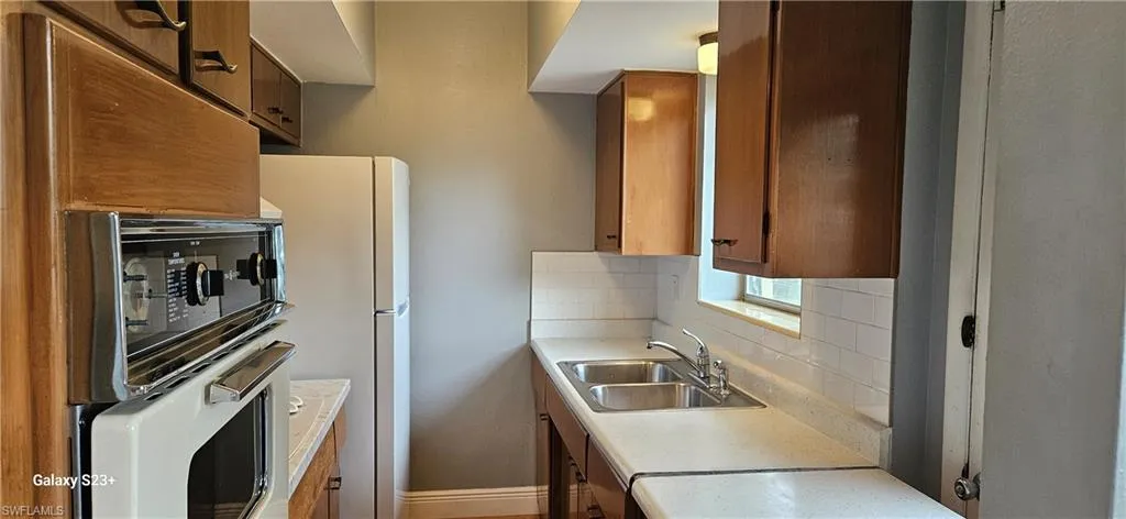 Kitchen featuring decorative backsplash, light countertops, freestanding refrigerator, and brown cabinetry