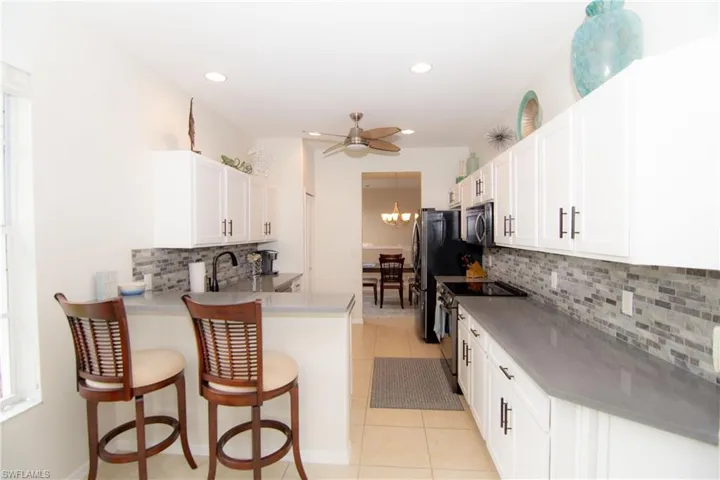 Kitchen featuring bright white Shaker style cabinetry with modern hardware, gray quartz countertops, stainless steel appliances, tile backsplash, recessed lighting and light tile floors.