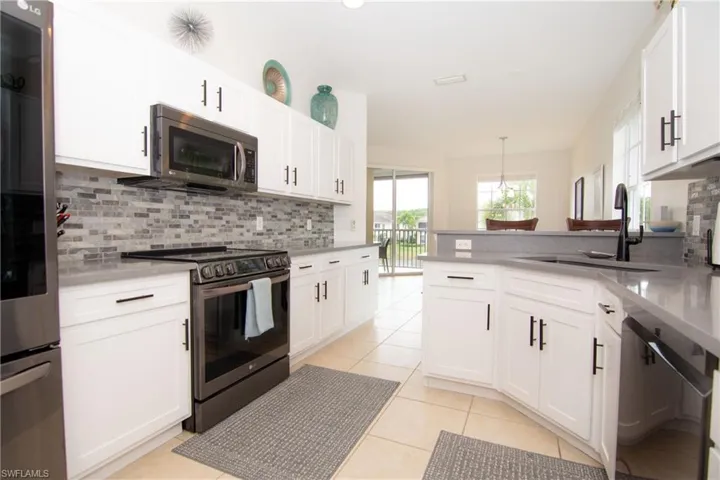 Kitchen featuring bright white Shaker style cabinetry with modern hardware, gray quartz countertops, stainless steel appliances, tile backsplash, recessed lighting and light tile floors.