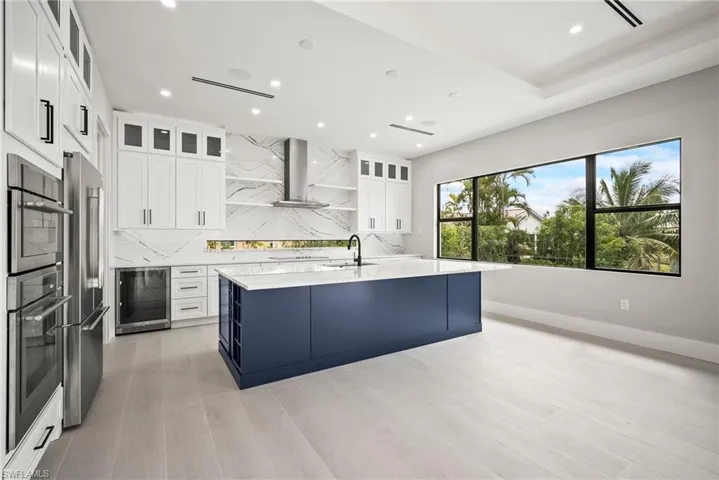 Kitchen with tasteful backsplash, white cabinetry, glass insert cabinets, wine cooler, and an island with sink
