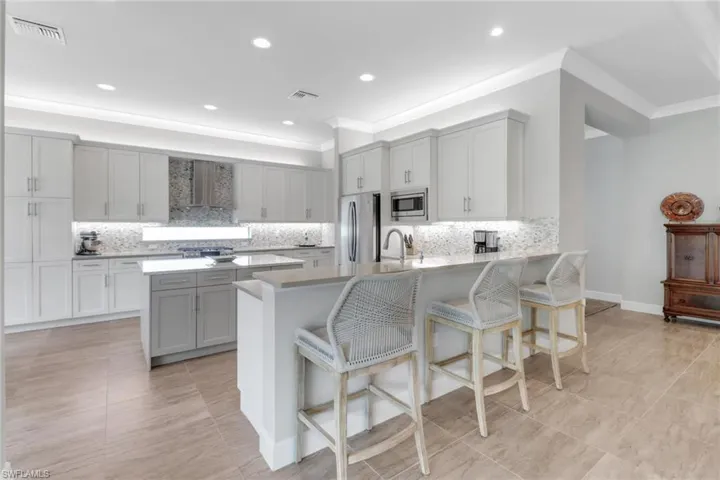 Kitchen with gray cabinetry, a peninsula, stainless steel appliances, wall chimney exhaust hood, and a kitchen bar