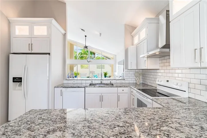 Kitchen featuring white appliances, glass insert cabinets, wall chimney exhaust hood, white cabinets, and vaulted ceiling