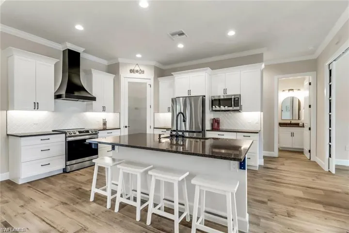 Kitchen with light wood-type flooring, an island with sink, white cabinetry, wall chimney exhaust hood, and appliances with stainless steel finishes