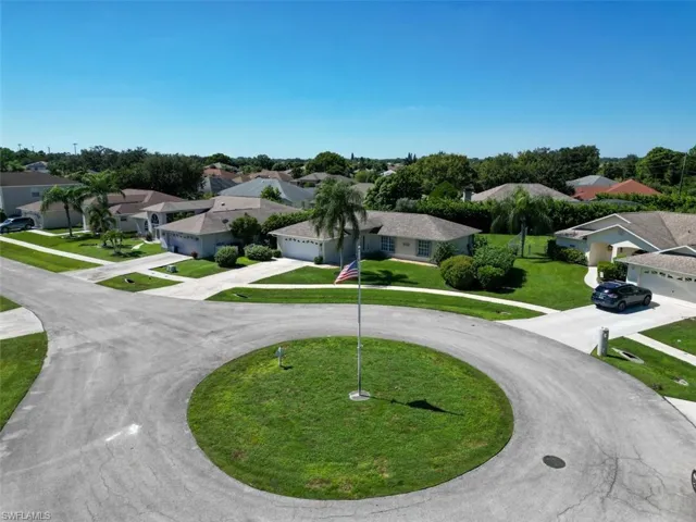 Aerial perspective of a residential street featuring paved cul-de-sac, central lawn area with flagpole, concrete driveways, and surrounding single-story residences with shingled roofing