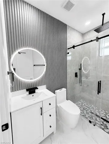 Modern bathroom featuring a single vanity with a white countertop and integrated sink, a backlit oval mirror, and a glass-enclosed shower with a hex tile floor