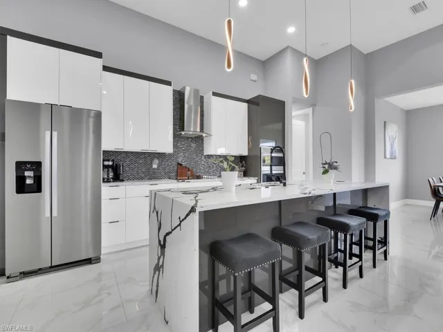 Kitchen with decorative light fixtures, an island with sink, wall chimney exhaust hood, stainless steel fridge, and white cabinets