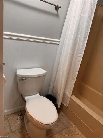 Full bath with toilet, shower / bath combo with shower curtain, stone finish flooring, and wainscoting