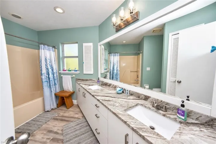 Bathroom featuring a dual-sink vanity with stone-patterned countertop, full-width wall mirror, recessed ceiling lighting, multi-light vanity fixture, and wood-finish flooring