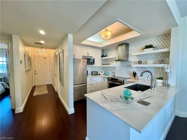 Kitchen with kitchen peninsula, stainless steel appliances, wall chimney exhaust hood, white cabinets, and dark wood-type flooring
