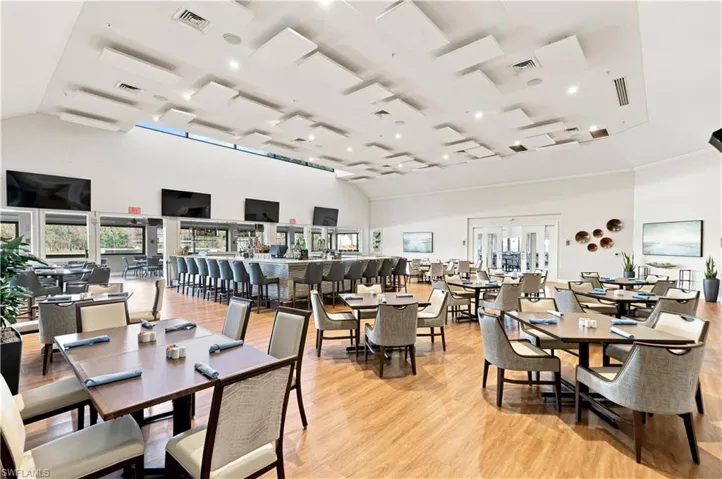 Spacious dining area featuring wood-finish flooring, a full-service bar with bar stools, multiple tables with seating, and high ceilings with decorative acoustic panels