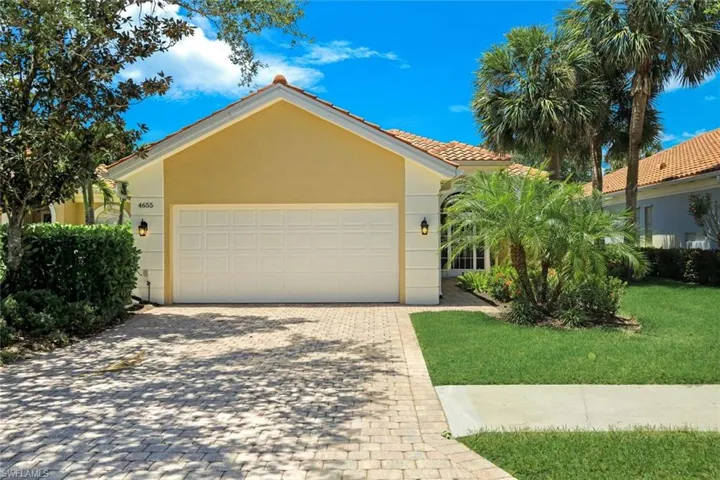 Stucco exterior with a tiled roof, featuring a paver driveway, an attached garage, and mature landscaping including palm trees