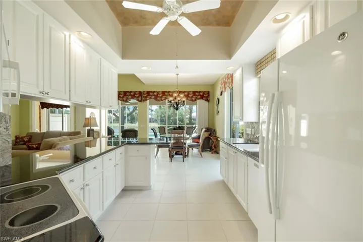 Bright kitchen featuring white cabinetry, dark countertops, white tile flooring, recessed lighting, and a tray ceiling with a ceiling fan