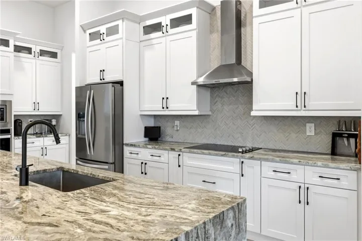 Kitchen with light stone countertops, wall chimney range hood, stainless steel appliances, glass insert cabinets, and white cabinetry