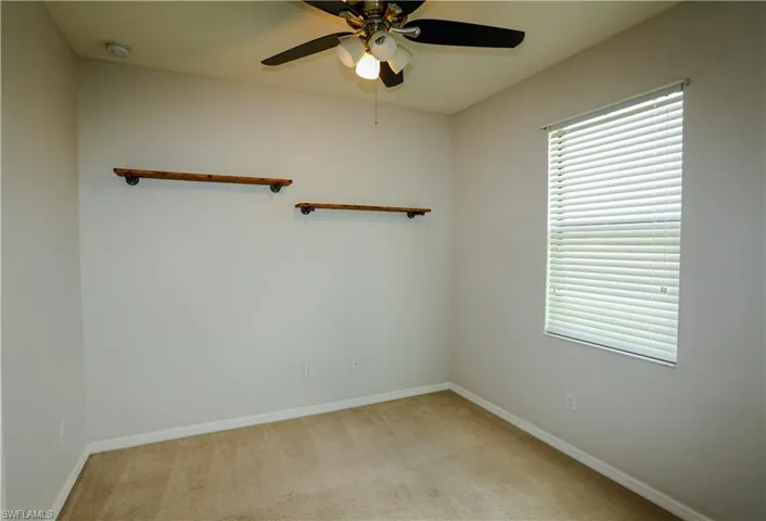 Neutral interior space featuring carpeted flooring, two wall-mounted wood shelves with metal piping brackets, a ceiling fan with integrated light fixture, and a window with horizontal blinds