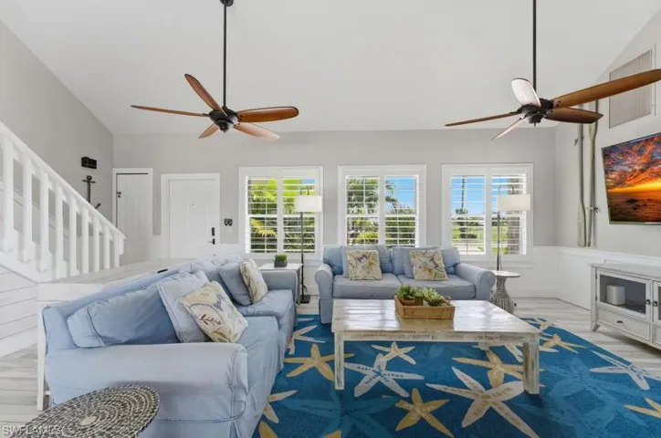 Spacious living area featuring vaulted ceilings, multiple windows with plantation shutters, wood-finish flooring, and a white staircase with a balustrade